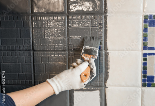 Repainting old dated kitchen ceramic tile back wall with modern gray chalk paint indoors at home. Giving old kitchen new look concept. Hand holding a paint brush tool. © FotoHelin