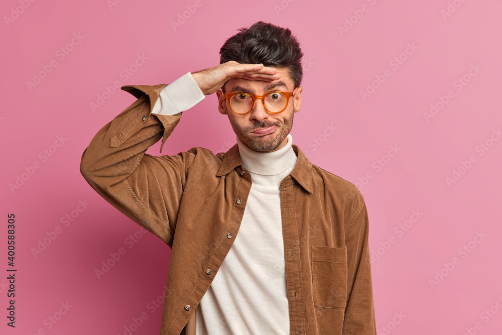 Waist up shot of puzzled displeased young man purses lips keeps hand near forehead tries to see something into distance wears spectacles turtleneck and brown shirt. Hmm what happening there. Waist up shot of puzzled displeased young man purses lips keeps hand near forehead tries to see something into distance wears spectacles turtleneck and brown shirt. Hmm what happening there.