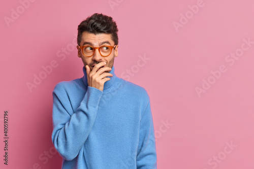 Studio shot of happy man tries to hide emotions giggles silently and covers mouth with hand looks aside positively dressed in casual clothes poses against pink background blank copy space on right