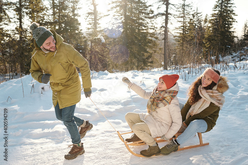 Two joyful girls in winterwear sitting on sledge while young man pulling them