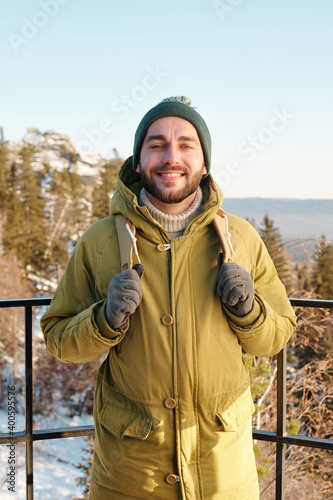 Young smiling male backpacker standing against trees and rocks covered with snow