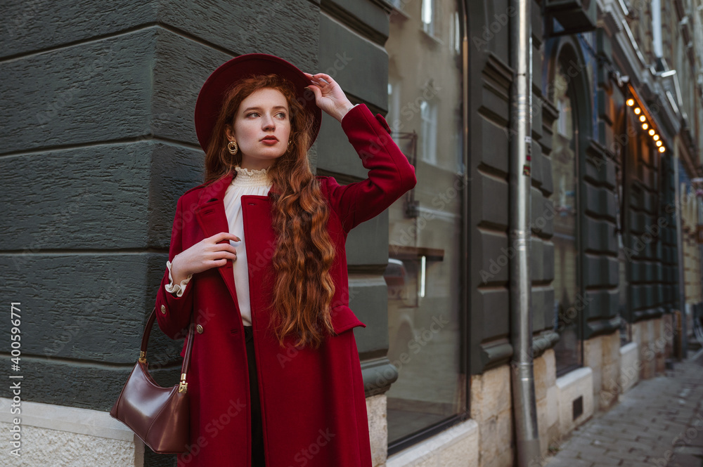 Autumn, winter fashion conception: elegant fashionable redhead woman wearing dark red fedora hat, stylish midi coat, holding small leather bag, posing in street of city.  Copy, empty  space for text