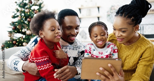 Close up of happy African American family at home sitting in decorated room near Christmas tree and using tablet Small cute kids sitting with mom and dad and tapping on tablet choosing presents online