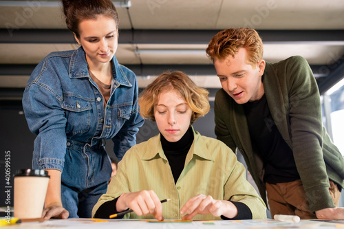 Blond serious girl sitting by desk while her two colleagues bending over table