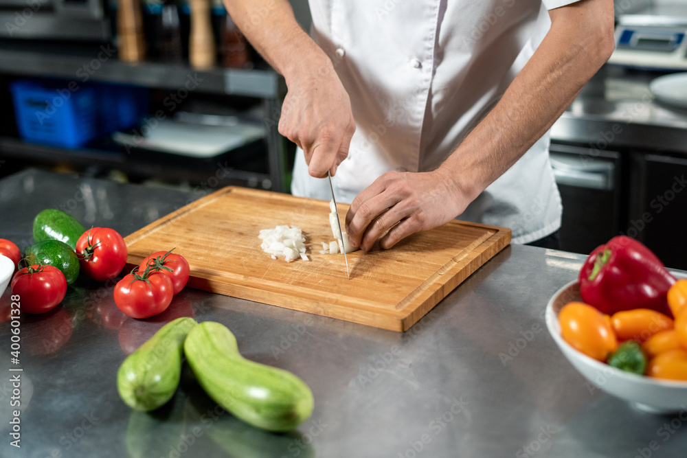 Hands of young male chef in uniform cutting fresh onions on board by table Hands of young male chef in uniform cutting fresh onions on board by table