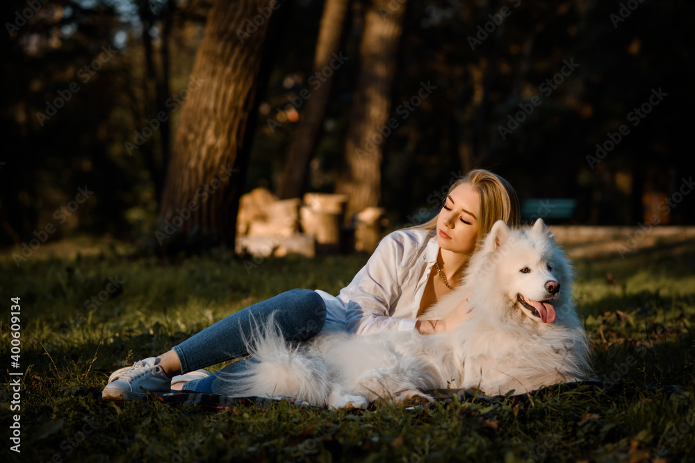beautiful woman in white shirt is hugging her white dog samoyed outdoors in the park and sitting on the grass. beautiful woman in white shirt is hugging her white dog samoyed outdoors in the park and sitting on the grass.