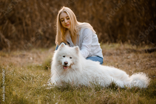 beautiful woman in white shirt is playing with her white dog samoyed outdoors in the park on the grass.