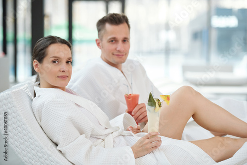 Young couple in bathrobes relaxing in spa center and having tropical cocktails