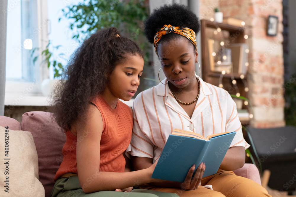 Young woman of African ethnicity and her cute teenage daughter reading book