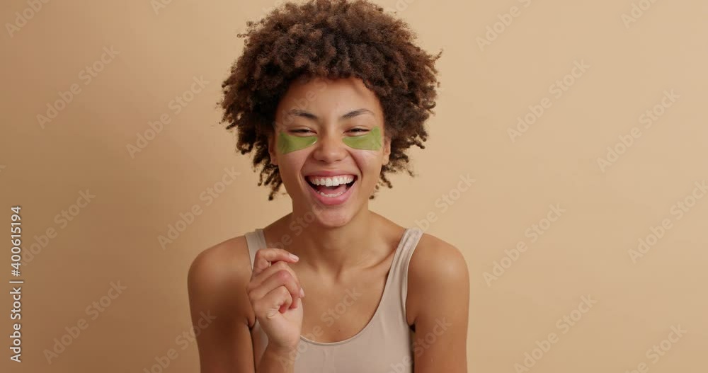 Overjoyed dark skinned young woman wears green patches under eyes to reduce dark circles and bags laughs out happily has fresh soft skin poses against brown background. Natural cosmetics concept Overjoyed dark skinned young woman wears green patches under eyes to reduce dark circles and bags laughs out happily has fresh soft skin poses against brown background. Natural cosmetics concept
