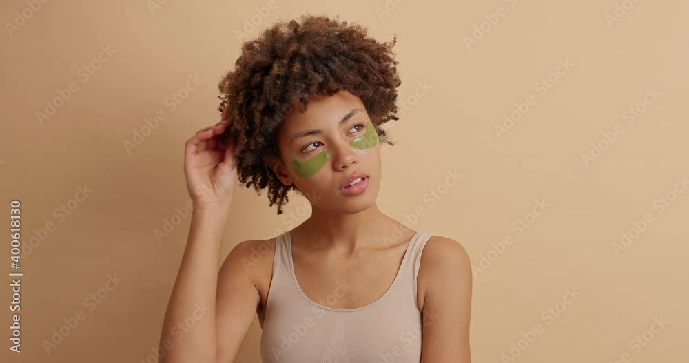 Thoughtful dark skinned young woman has natural makeup and applies collagen patches under eyes on fresh skin curls hair contemplates about somethig isolated over brown background. Beauty concept Thoughtful dark skinned young woman has natural makeup and applies collagen patches under eyes on fresh skin curls hair contemplates about somethig isolated over brown background. Beauty concept