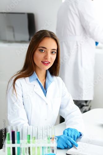 Fototapeta Naklejka Na Ścianę i Meble -  Smiling beautiful technician woman portrait and sample bottle with poison fluid. Medical worker in uniform use reagent tube for virus infection exam or biological toxic reaction, drug creation