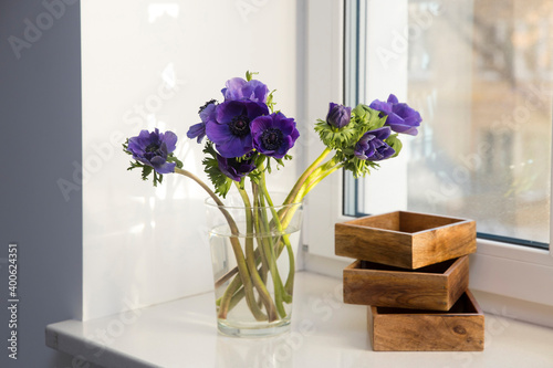 A bouquet of blue anemones in a glass vase next to three wooden boxes for storing small items on the windowsill