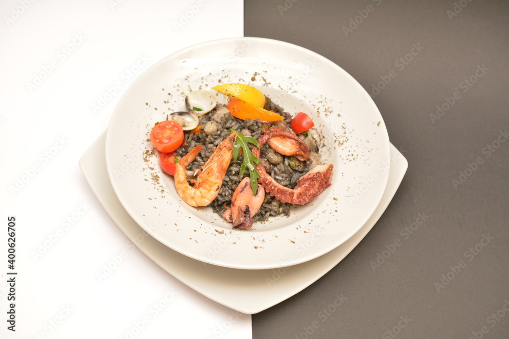 Risotto with seafood and cuttlefish ink on a white plate. Decorated with boiled shrimp and lemon wedges. Located on a black and white background. Can be used in the restaurant menu.