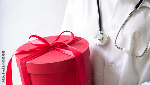 Concept for christmas and new year medical banner.Female doctor in white coat,stethoscope,latex gloves holds big red gift box with bow.Gift for birthday,doctor's day,March 8,mother's day.Copy space