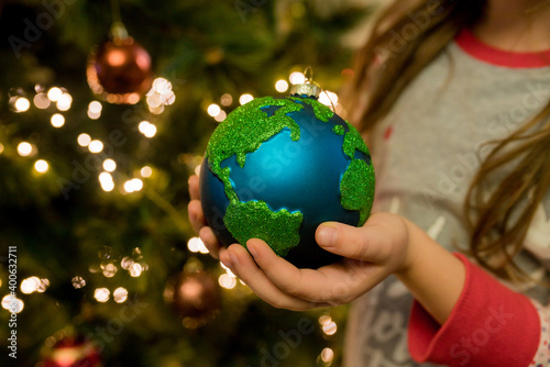 Fototapeta Naklejka Na Ścianę i Meble -  Close-up of little girl hanging Xmas glass bauble decoration ornament globe planet earth on christmas tree. Merry christmas and new year concept. Selective focus