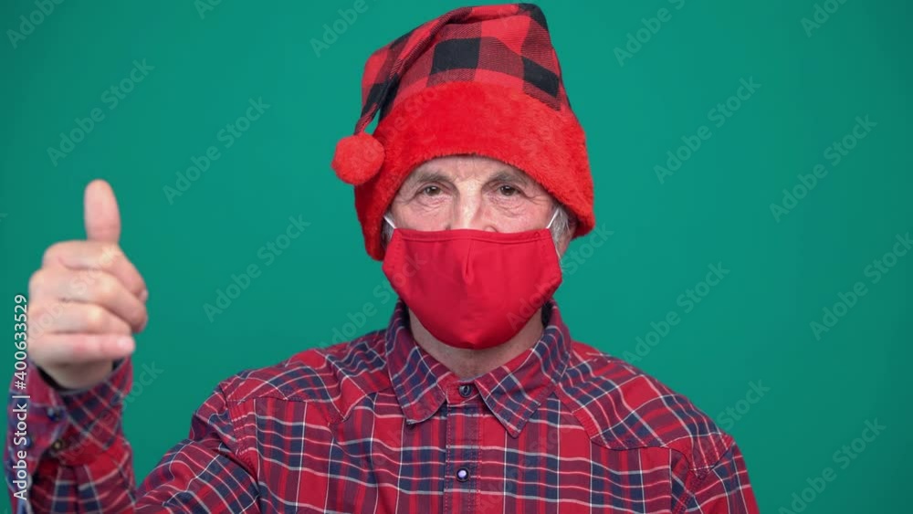 Portrait of old man wearing Christmas red hat and medical protective face mask showing thumbs up and approval sign with hand Portrait of old man wearing Christmas red hat and medical protective face mask showing thumbs up and approval sign with hand