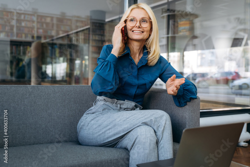 Elegant business woman sit at the couch in office, smiling. Senior lady using smartphone, communicating with colleagues, discuss new project. Middle aged female employee working distantly