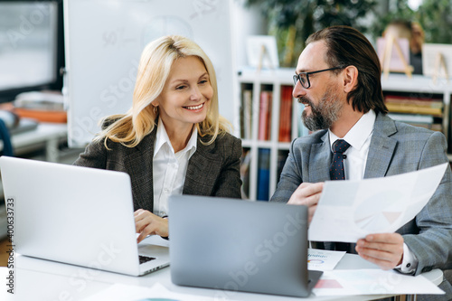 Friendly mature smiling colleagues at workplace. Business partners brainstorming together, discussing new project. Senior office employees looking at financial graphs while sitting at the work desk