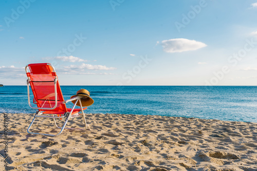 Fototapeta Naklejka Na Ścianę i Meble -  straw hat on top of a red chair on a deserted beach