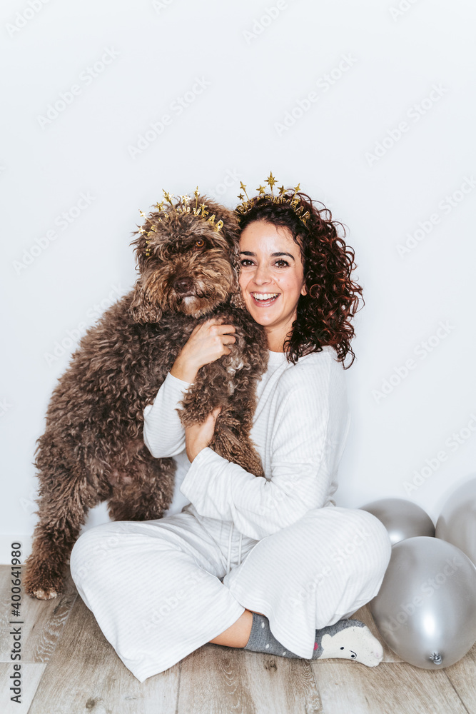 Beautiful brunette woman in pajamas hugging her dog and both looking at the camera in the New Year's party at home. They are wearing Christmas headbands. New Year's Eve party at home concept