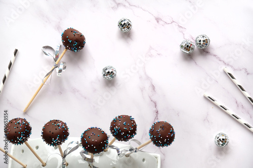 Yummy cake pops covered with chocolate and colored sugar icing. Party decorations, drink paper straws and disco balls on white marble background. Merry Christmas and a Happy New Year