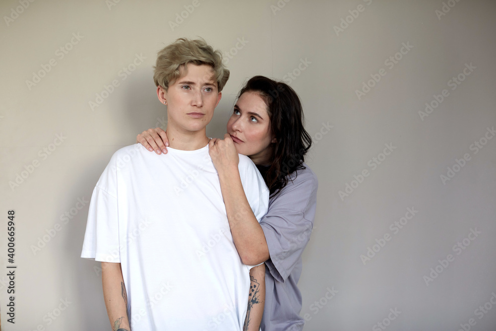 Young brunette embracing and looking at androgynous woman with short hair against gray wall Young brunette embracing and looking at androgynous woman with short hair against gray wall