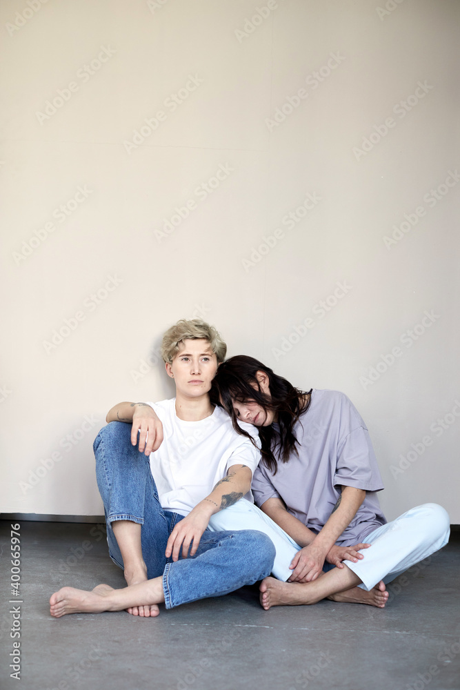 Young barefoot woman leaning on shoulder of girlfriend with short hair while relaxing against gray wall together Young barefoot woman leaning on shoulder of girlfriend with short hair while relaxing against gray wall together