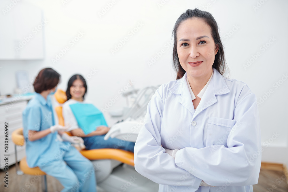 Portrait of confident smiling female Vietnamese dentist folding arms and looking at camera, her assistant talking to patient in background Portrait of confident smiling female Vietnamese dentist folding arms and looking at camera, her assistant talking to patient in background