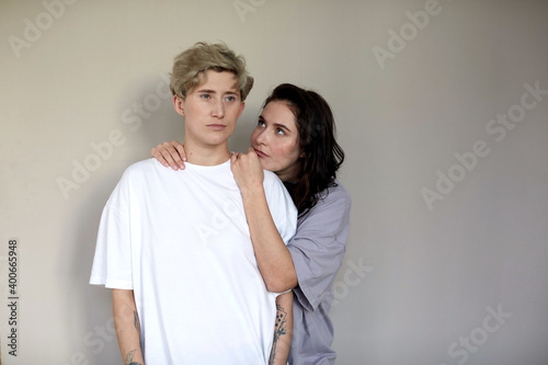 Young brunette embracing and looking at androgynous woman with short hair against gray wall