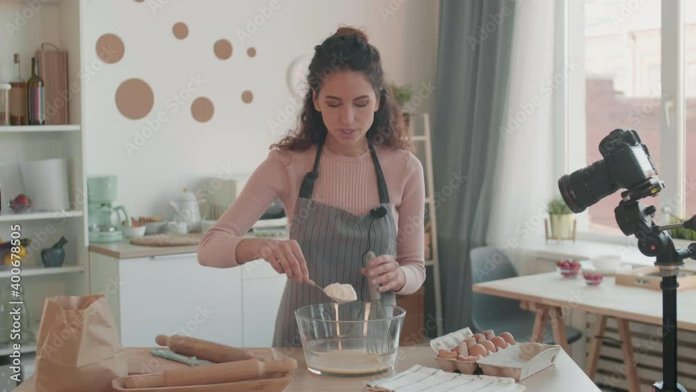 Medium POV of smiling Caucasian young woman wearing apron with microphone clipped on it, standing by kitchen table, adding flour to dough, smiling, talking, looking on camera, another one filming bowl Medium POV of smiling Caucasian young woman wearing apron with microphone clipped on it, standing by kitchen table, adding flour to dough, smiling, talking, looking on camera, another one filming bowl