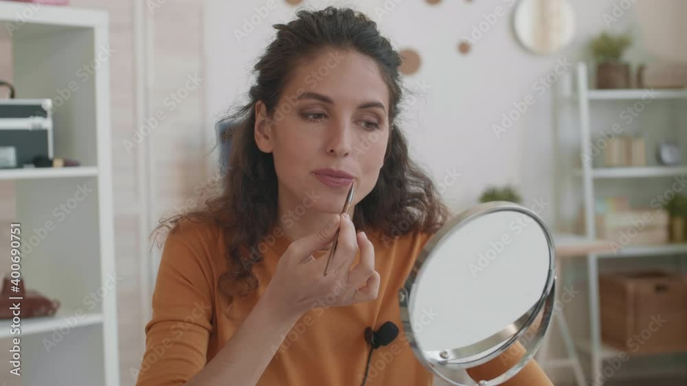 Close-up POV of young Caucasian female beauty vlogger looking in table mirror, applying makeup on lips with brush, then removing excess by biting facial tissue paper Close-up POV of young Caucasian female beauty vlogger looking in table mirror, applying makeup on lips with brush, then removing excess by biting facial tissue paper