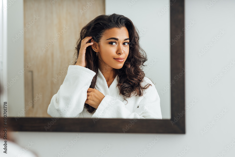 Beautiful female wearing bathrobe looking at a mirror and touching her hair Beautiful female wearing bathrobe looking at a mirror and touching her hair