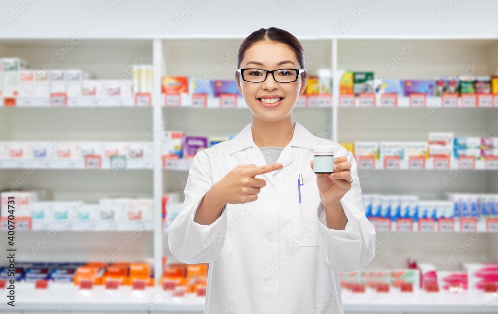 medicine, profession and healthcare concept - happy smiling asian female pharmacist or doctor holding jar of pills over pharmacy background