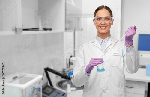 science  chemistry and people concept - young female scientist in goggles with pipette chemical in flask making test or research over laboratory background