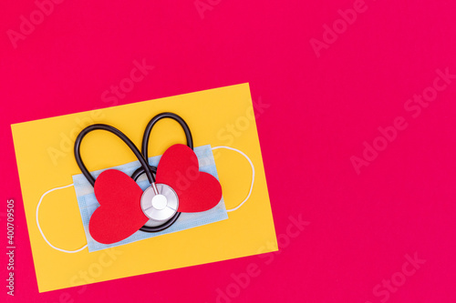 Black heart shaped stethoscope on medical protective mask and red two paper heart shape on trendy yellow Illuminating and red background, top view, copy space. Health care and cardiology concept