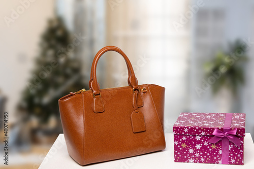 Template for christmas sale. Elegant brown female luxury handbag and a gift box on table against blurred xmas tree background indoor. Advertising fashionable woman accessories.