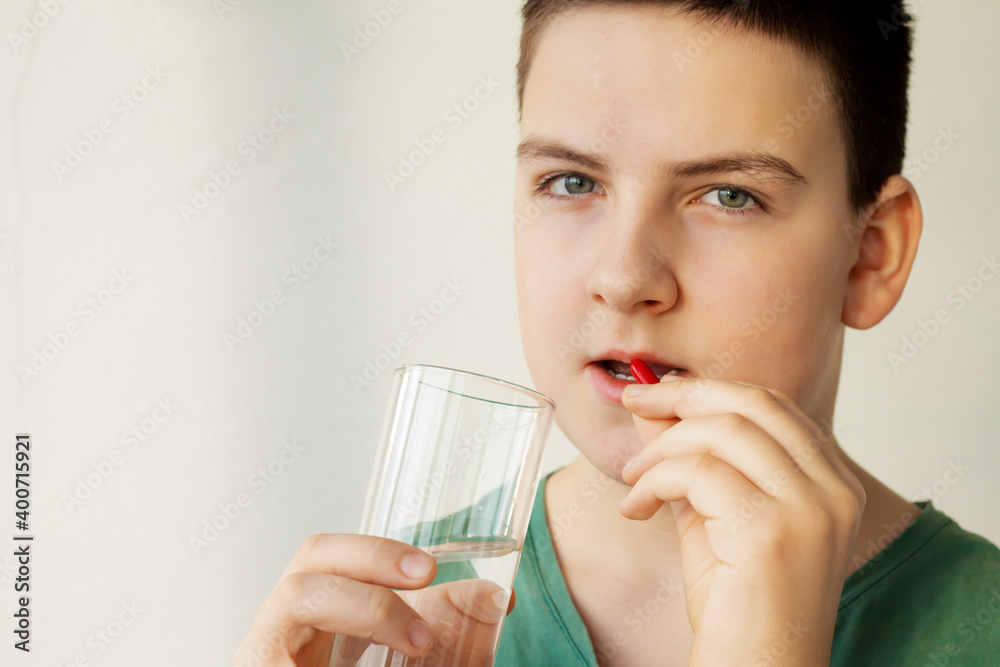 a boy (young man) holding a red capsule with medicine and a glass of water; health care concept, additives or vitamins, disease treatment a boy (young man) holding a red capsule with medicine and a glass of water; health care concept, additives or vitamins, disease treatment