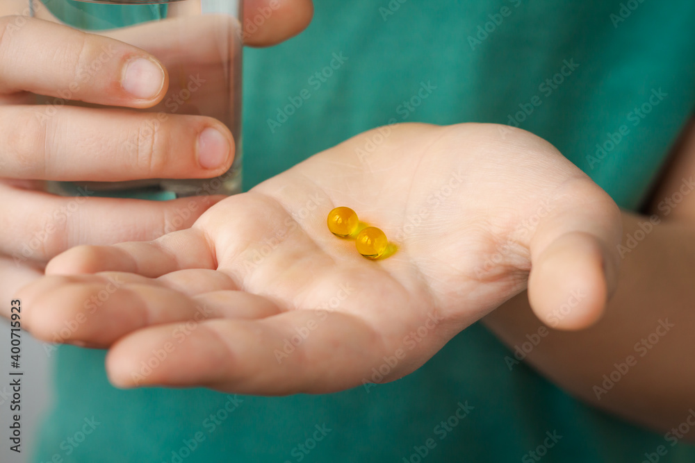 two yellow medicine capsules with glass of water closeup, fish oil round yellow pills on man hand, sport nutritional supplements, disease treatment, medicine additives or vitamins