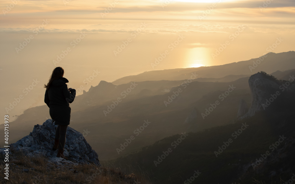 Fototapeta premium A girl stands on a mountain and looks at the sunset. Atmospheric evening view of the mountains and the sea. beautiful aerial perspective. The concept of relaxation and meditation. Bright sunlight.