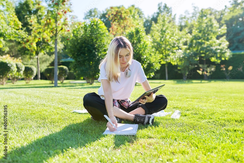 Teenage student in headphones with digital tablet sitting on lawn writing in notebook Teenage student in headphones with digital tablet sitting on lawn writing in notebook