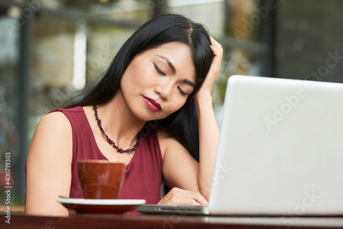 Tired young businesswoman almost sleeping when sitting at cafe table with cup of coffee and laptop