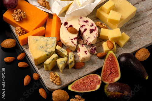 Cheese plate. Cheddar, Brie, Red Leicester, Wensleydale with sweetened dried cranberries, Blue Stilton, almond and walnut on a marble board with grape and figs on a black background.