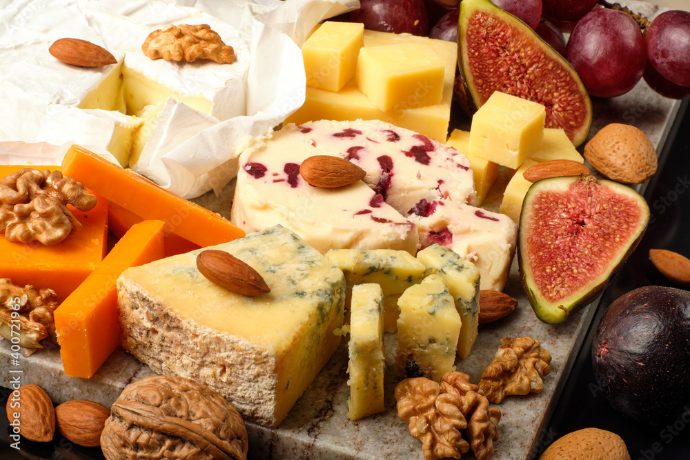 Cheese plate. Cheddar, Brie, Red Leicester, Wensleydale with sweetened dried cranberries, Blue Stilton, almond and walnut on a marble board with grape and figs on a black background. Cheese plate. Cheddar, Brie, Red Leicester, Wensleydale with sweetened dried cranberries, Blue Stilton, almond and walnut on a marble board with grape and figs on a black background.