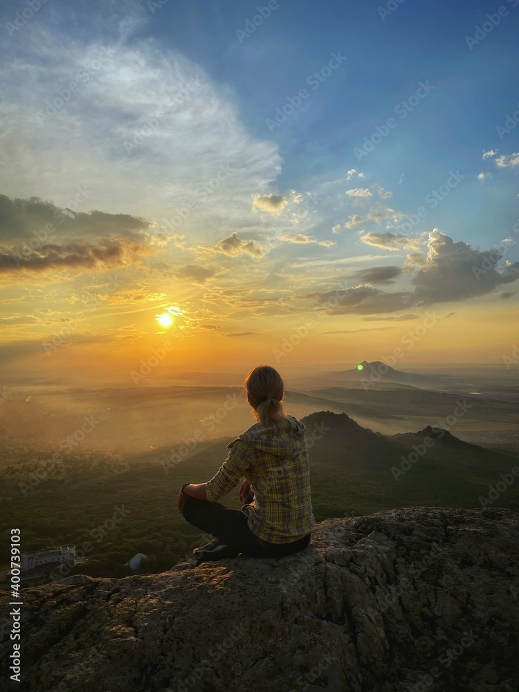 The girl sits with her back on a rock and looks at the sunset in the mountains. Beautiful clouds all around