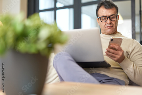 Work in your comfort zone. Handsome latin middle aged businessman in eyeglasses using smartphone while working on the laptop from home