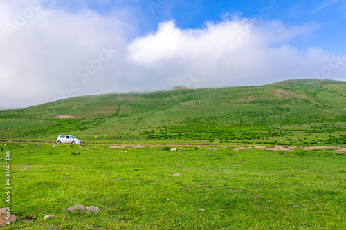 White car drives on caucasian green wilderness with sky and meadow view. Copy paste road trip background