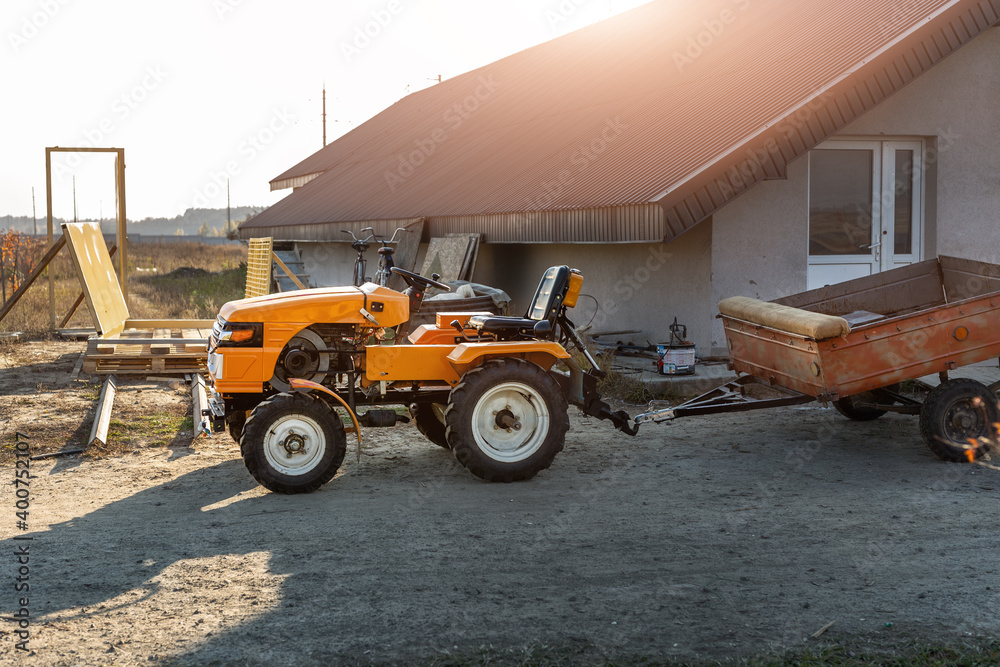 Obraz premium Small mini red modern orange new tractor with trailer standing near hangar building at farm countryside during sunset or sunrise. Small agricultural machinery. Rural country farmland scene background