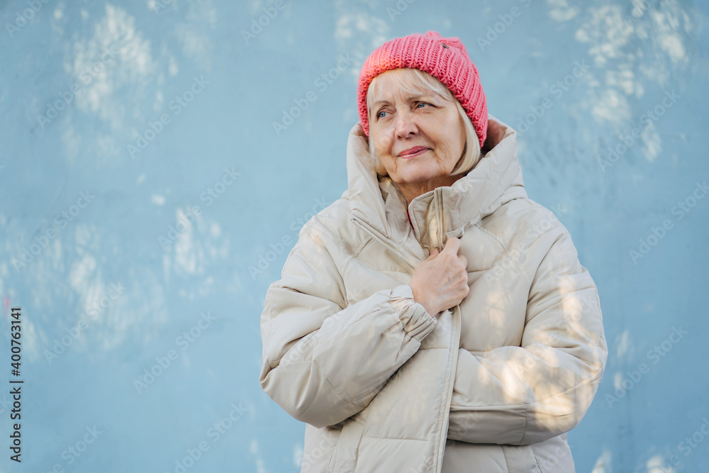Senior female in trendy outerwear. Aged woman in knitted pink hat smiling and adjusting warm coat looking away against light blue wall on street Senior female in trendy outerwear. Aged woman in knitted pink hat smiling and adjusting warm coat looking away against light blue wall on street