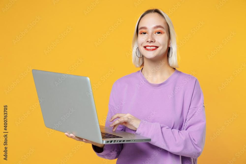 Naklejka premium Smiling young blonde caucasian woman 20s with bob haircut bright makeup wearing basic purple shirt using modern laptop pc computer typing keyboard isolated on yellow color background studio portrait.